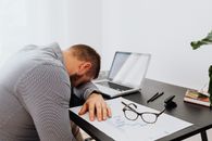 A man in a gray shirt sits tiredly at a desk with a laptop and documents, showing signs of workplace stress.
